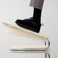 On-foot beeld van UGG Lowmel Lo Black met zwarte sokken, voet rustend op modern designstoel in minimalistische studio