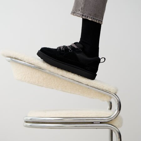 On-foot beeld van UGG Lowmel Lo Black met zwarte sokken, voet rustend op modern designstoel in minimalistische studio