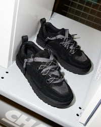 Flatlay van twee zwarte UGG Lowmel Lo sneakers in witte kast, met zicht op veters, suède textuur en rubberen zool