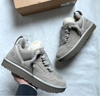 Close-up van twee UGG Lowmel sneakers in pumice kleur in een hand, met de schoendoos zichtbaar op de achtergrond