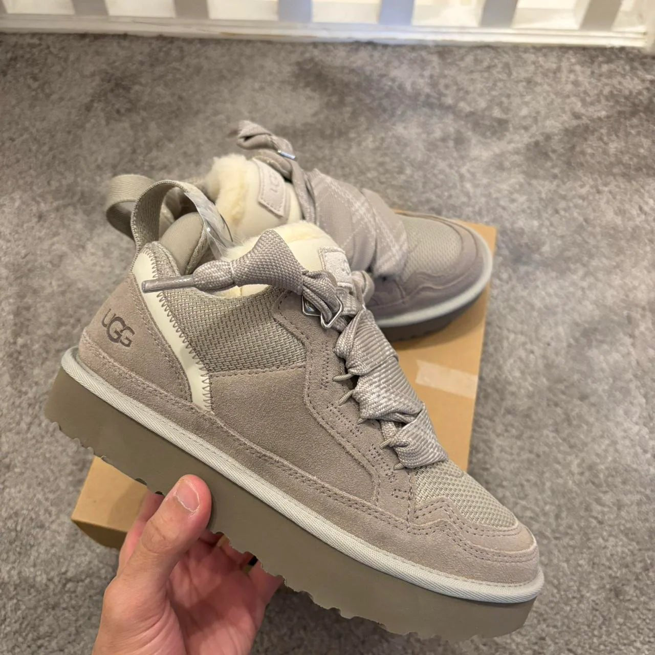 Hand houdt één UGG Lowmel Pumice vast boven een grijze vloer met subtiel tapijtpatroon, schoen steunt op de doos