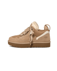 Zijkant van de UGG Lowmel Sand voor kinderen, beige upper met shearling tong en robuuste bruine zool tegen witte achtergrond