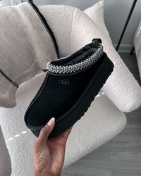 Close-up van UGG Tazz Slipper Black 1122553-BLK in hand, met zichtbare suède textuur, heritage braid afwerking en stevige platformzool, gefotografeerd door Snrkickz.