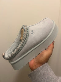 Close-up van UGG Tazz Slipper Goose 1122553-GOO, lichtgrijs suède met gevlochten rand en stevige platformzool, vastgehouden voor detailweergave door Snrkickz.